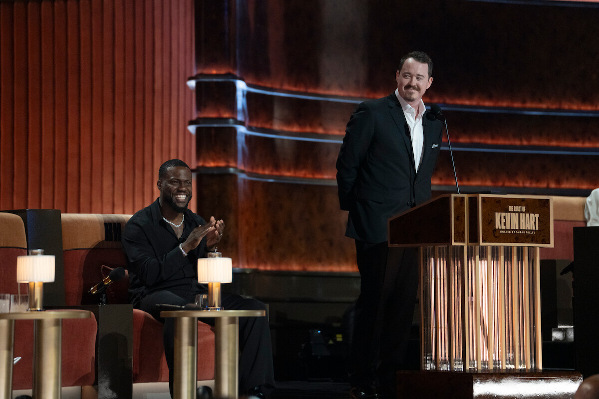 Two men on stage at a comedy roast, one sitting and clapping, the other standing and speaking at a podium labeled "Kevin Hart." The setting has warm lighting with elegant furniture and a formal, theatrical backdrop.