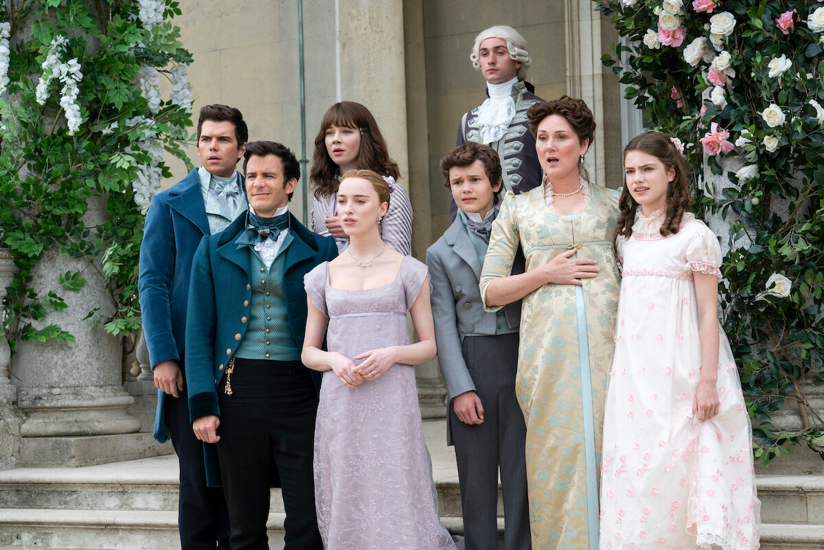 A group of men and women in Regency-era costumes stand outside a grand stone building adorned with flowers, posing on steps, suggesting a formal gathering or family portrait in a historical setting.