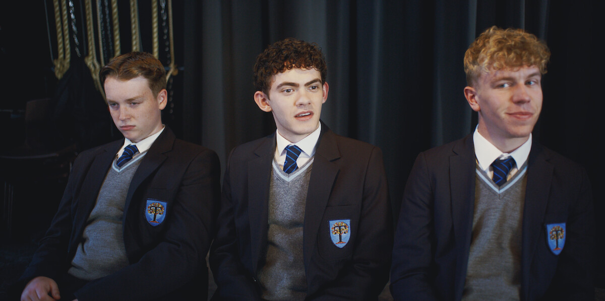 Three male students in school uniforms sit together on a bench indoors, with dark curtains and ropes in the background, appearing to be in a formal or staged setting, each displaying different facial expressions.