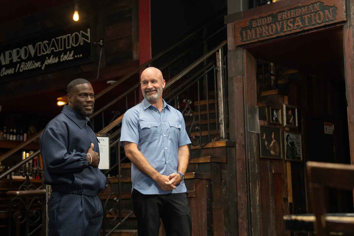 Two men stand smiling inside a comedy club with rustic décor, wood walls, and stairs. Signs read "IMPROVISATION" and "BUDD FRIEDMAN IMPROVISATION" above the entrance. Warm ambient lighting creates a welcoming setting.