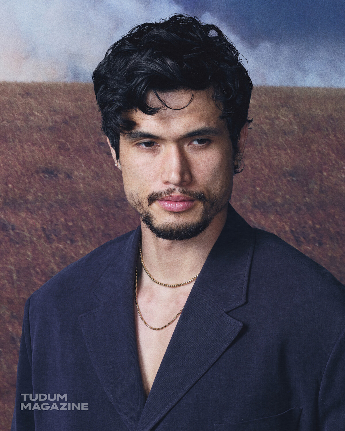 A man with dark hair and a beard wearing a navy blazer and gold chains stands in front of a blurred outdoor landscape with cloudy sky.