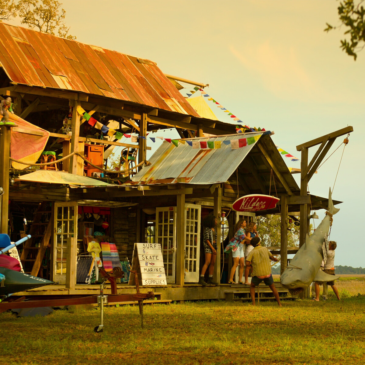 A group of people gathers at a rustic wooden shack decorated with flags, with surfboards, a kayak, and a large hanging fish outside, set in a grassy outdoor environment near water at sunset.
