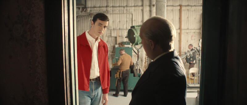 A young man in a red jacket stands in a doorway looking at an older man in a suit, with a busy workshop or studio visible in the background.