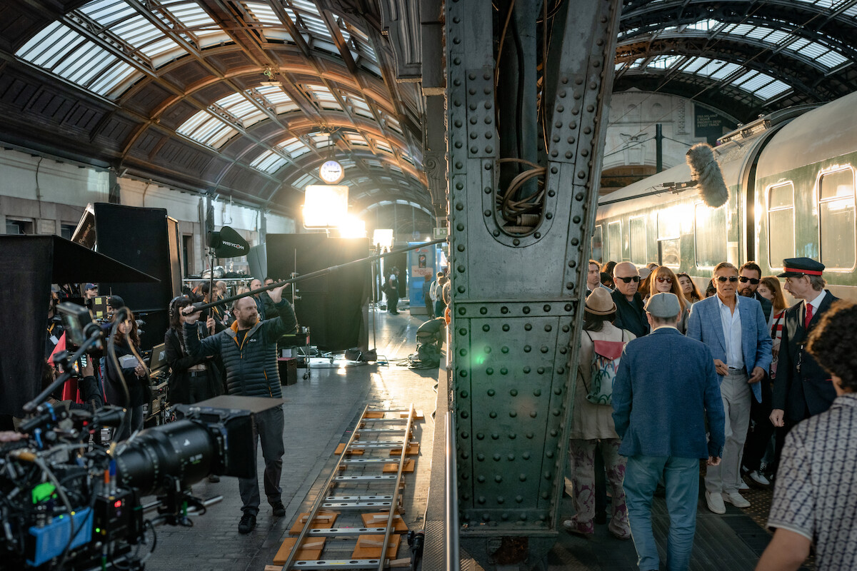 Film crew shooting a scene at a vintage train station set, with actors in costume, camera equipment, lighting, and crew members visible. The mood is busy and professional, capturing a behind-the-scenes atmosphere.