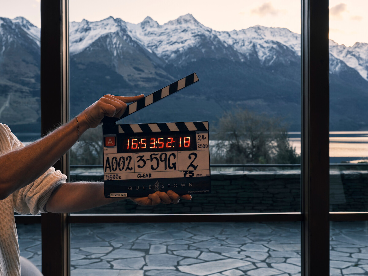 A person holds a film clapperboard indoors in front of large windows, with a scenic view of a lake, stone terrace, and snow-capped mountains in the background during daylight.