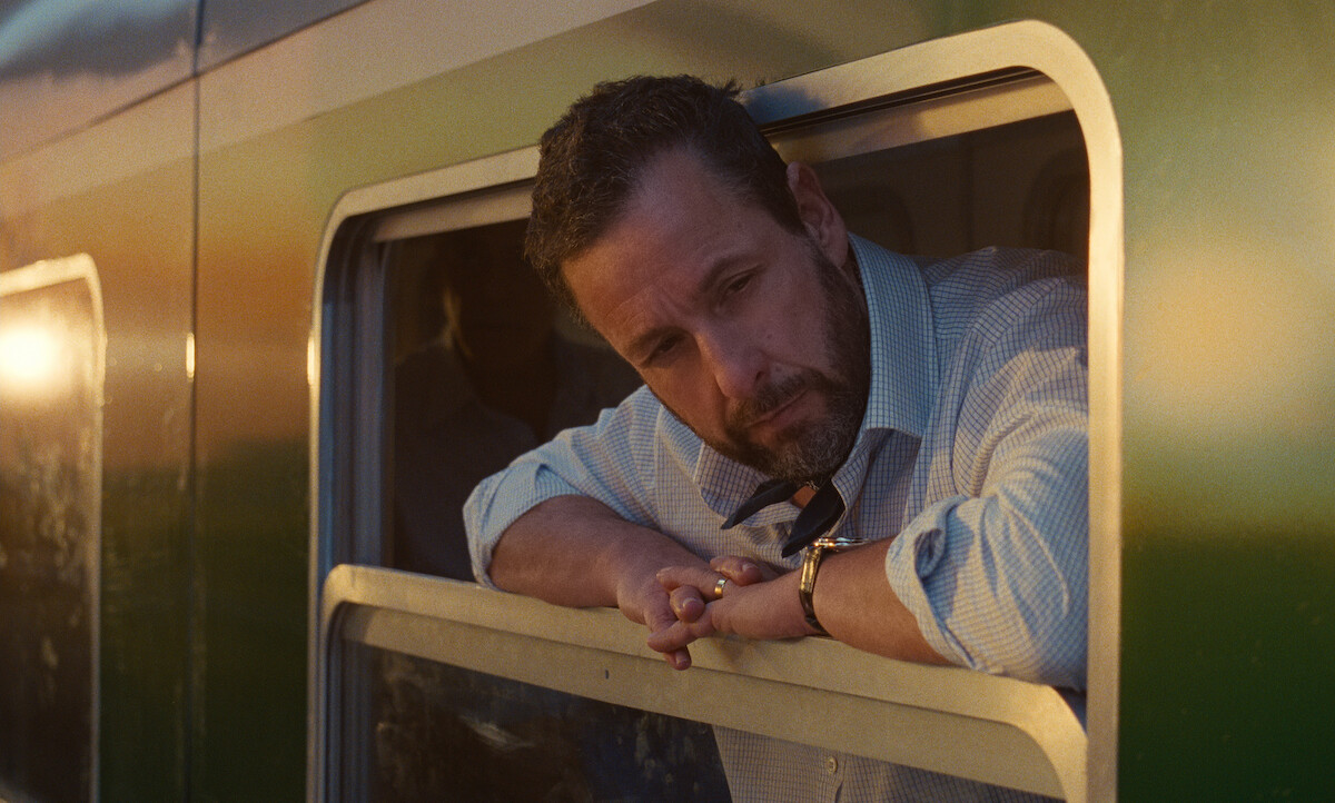 A man leans out of a train window at sunset, gazing thoughtfully outside. The environment suggests a peaceful, reflective mood inside a green train carriage.