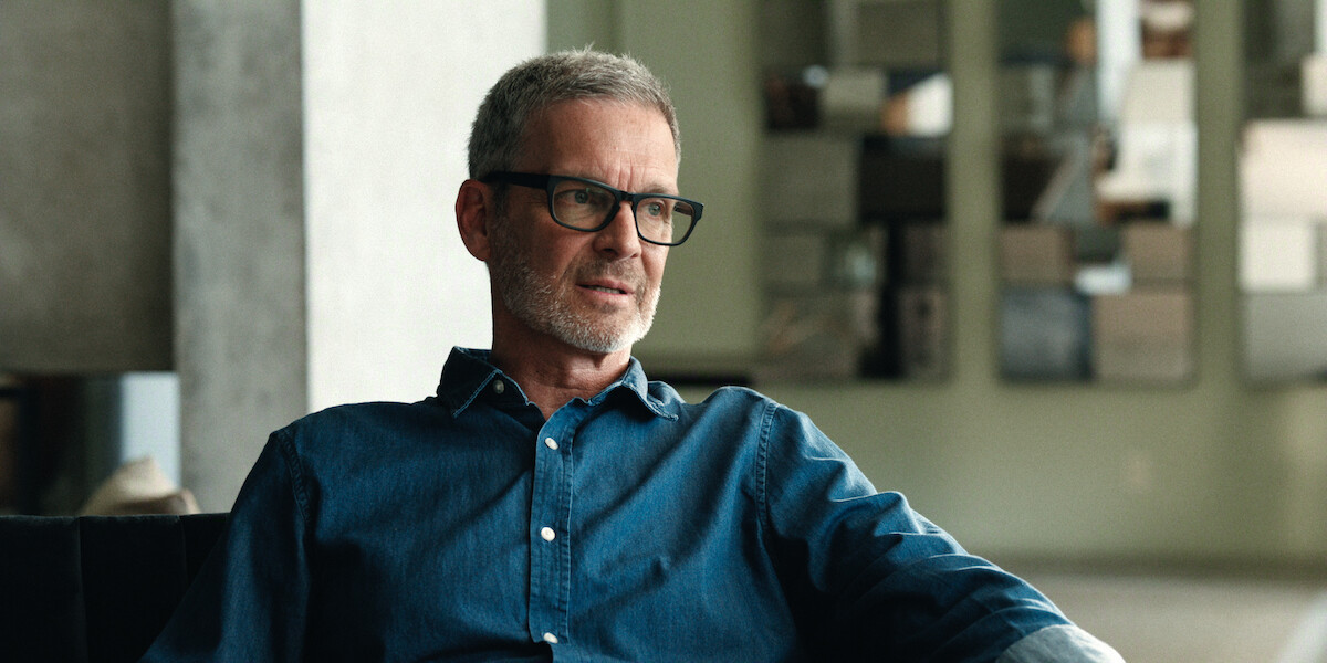 Man with gray hair and glasses in a denim shirt sitting indoors on a sofa, with a contemporary interior featuring neutral colors and modern wall art in the background.