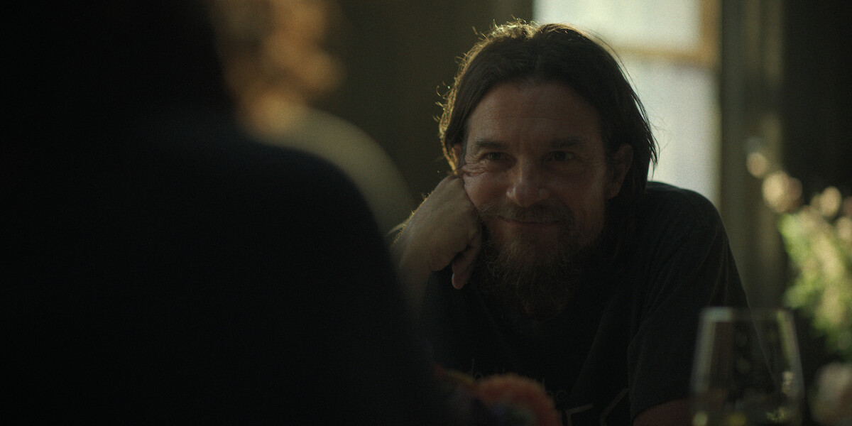 Man with long hair and beard smiling and resting his chin on his hand, sitting at a table indoors with soft natural light and blurred background, in a relaxed and intimate setting.