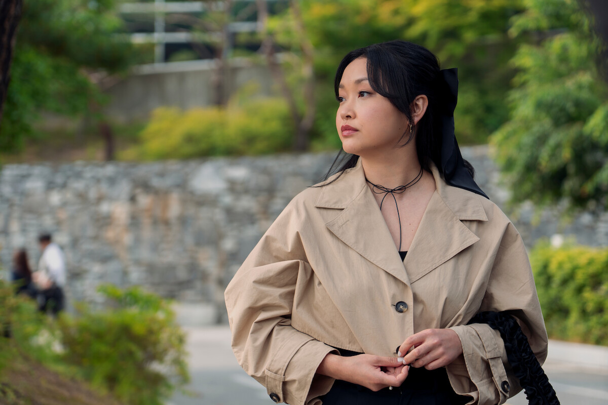Woman in beige coat standing outdoors in a park-like setting with green trees, stone wall, and soft natural light, looking to the side thoughtfully.