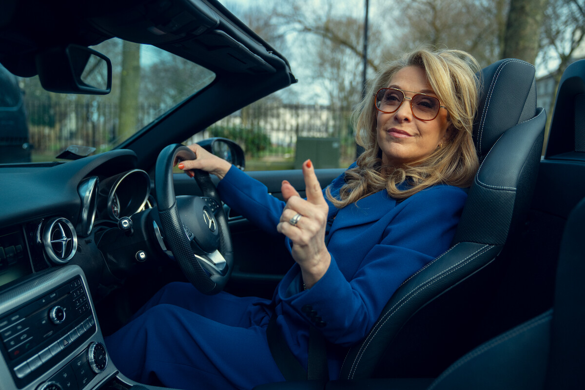 Confident woman in blue suit and sunglasses sitting in a convertible car, hand on steering wheel, pointing with a relaxed expression. The setting is outdoors with trees and muted daylight, creating a calm, empowered mood.