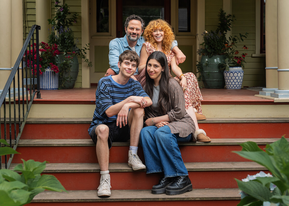 Four people sit smiling together on the steps of a porch in front of a house, surrounded by potted plants and greenery, creating a warm and inviting family portrait.