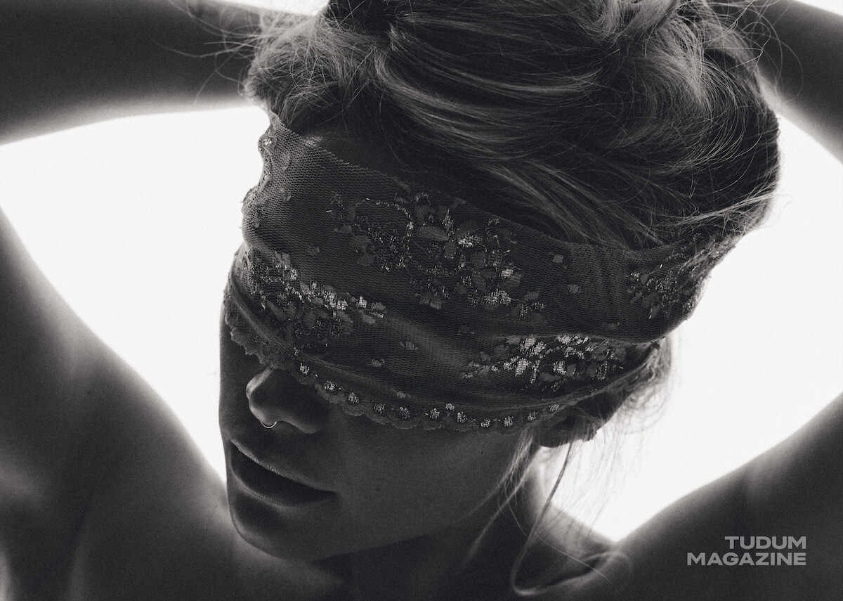 A person in a close-up, black and white photo with a decorative lace blindfold, arms raised behind their head, light background, studio setting, and “TUDUM MAGAZINE” watermark in the corner.
