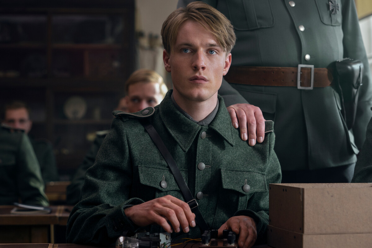 Werner Pfennig (Louis Hofmann), a teenage boy with light features, dirty-blonde hair and blue eyes sits at a desk in a military uniform. A military officer has his hand on Werner’s shoulder and there are other young military recruits in the background. On the desk in front of Werner sits an assembled radio and cardboard box.