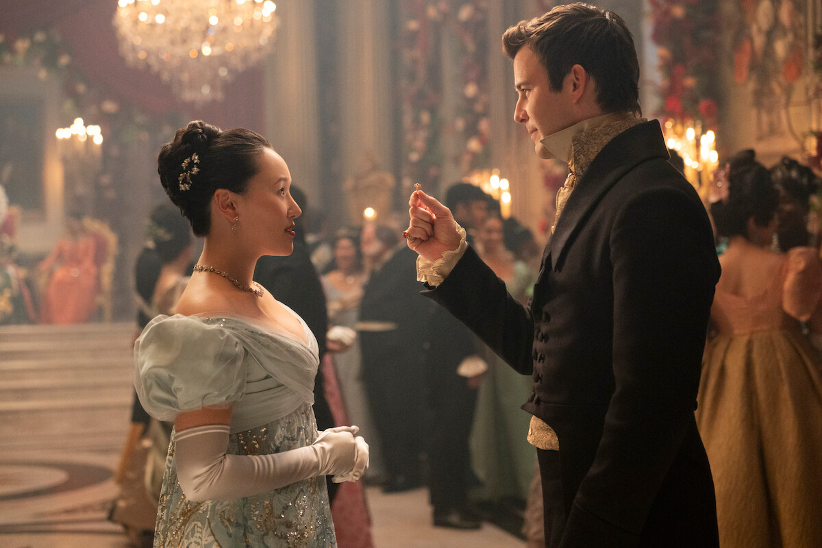 A woman and a man in elegant period costumes converse at a formal ballroom event, surrounded by other guests, chandeliers, and elaborate decorations.