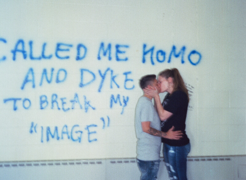 Students kissing in front of a graffiti wall that reads" Called me homo and dyke to break my image."