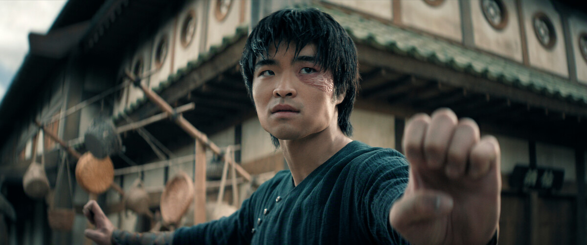 Dallas Liu as Zuko with a scarred face stands in a martial arts pose on a traditional village street, focused and ready to fight, with old wooden buildings and hanging baskets in the background.