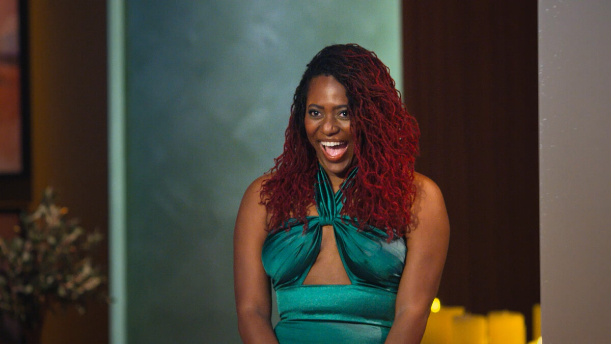 Woman with long curly red hair wearing a green dress, smiling in a warmly lit indoor setting with decorative background elements.