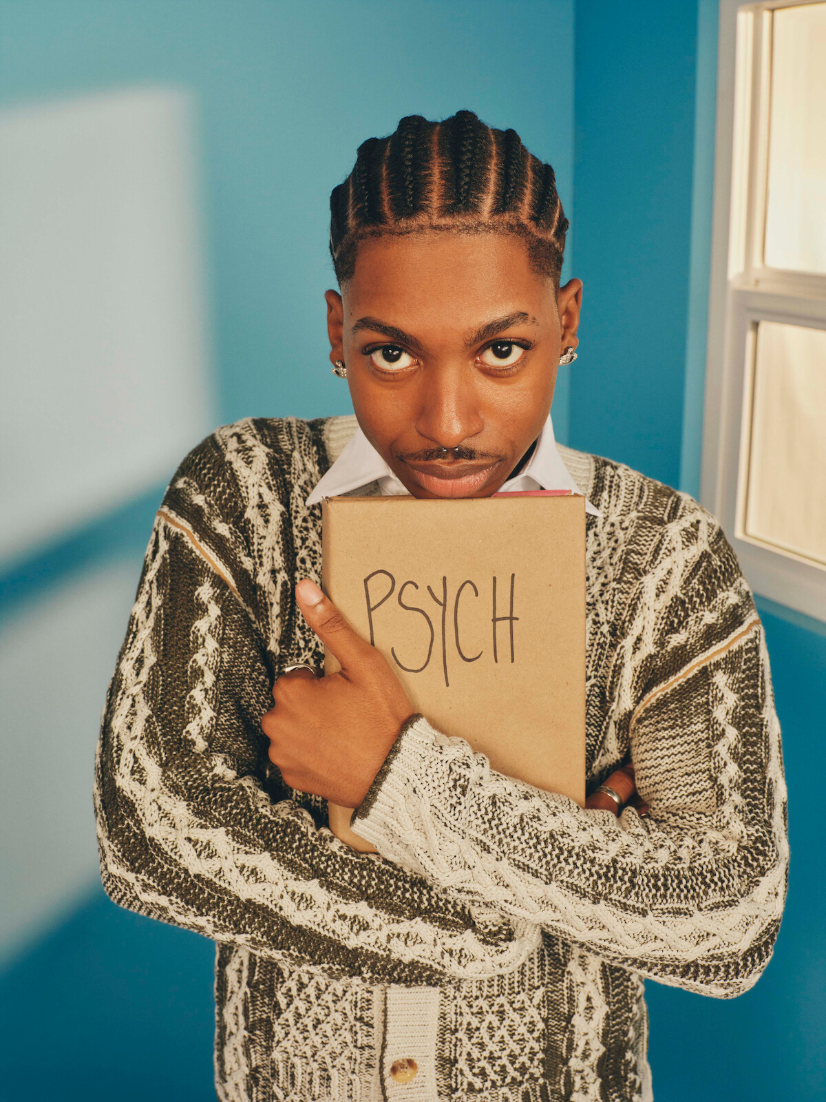 A young person with braids, wearing a patterned sweater, stands in a blue room holding a notebook labeled "PSYCH" to their chest, looking directly at the camera near a window with sunlight.