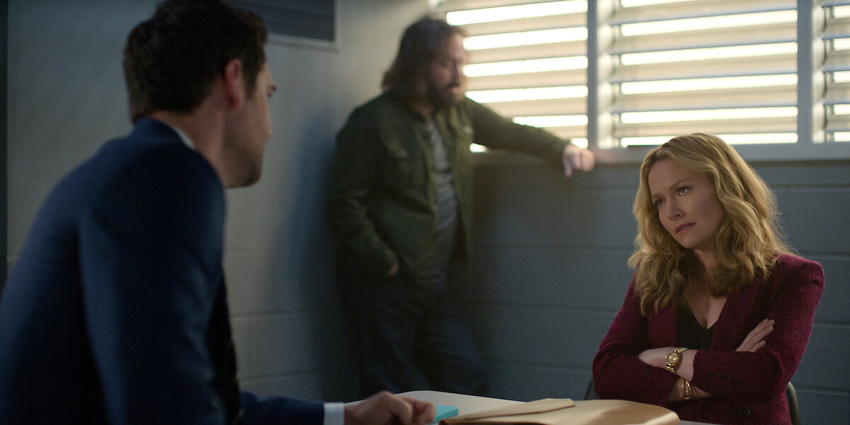 A woman sits at a table with arms crossed, looking serious, while a man in a suit sits across from her and another man stands in the background. The setting appears to be an interrogation room with barred windows.