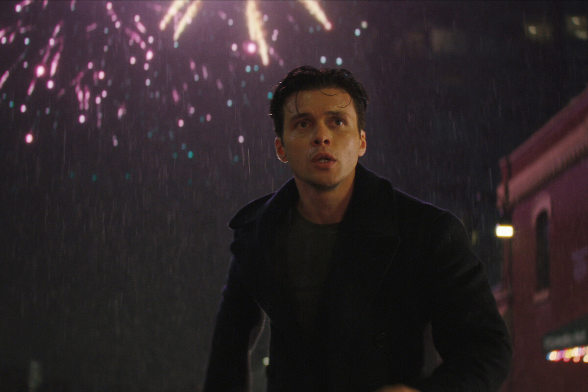 A young man in a dark coat stands outside at night in the rain, looking up as colorful fireworks explode in the sky above a city street with buildings in the background.
