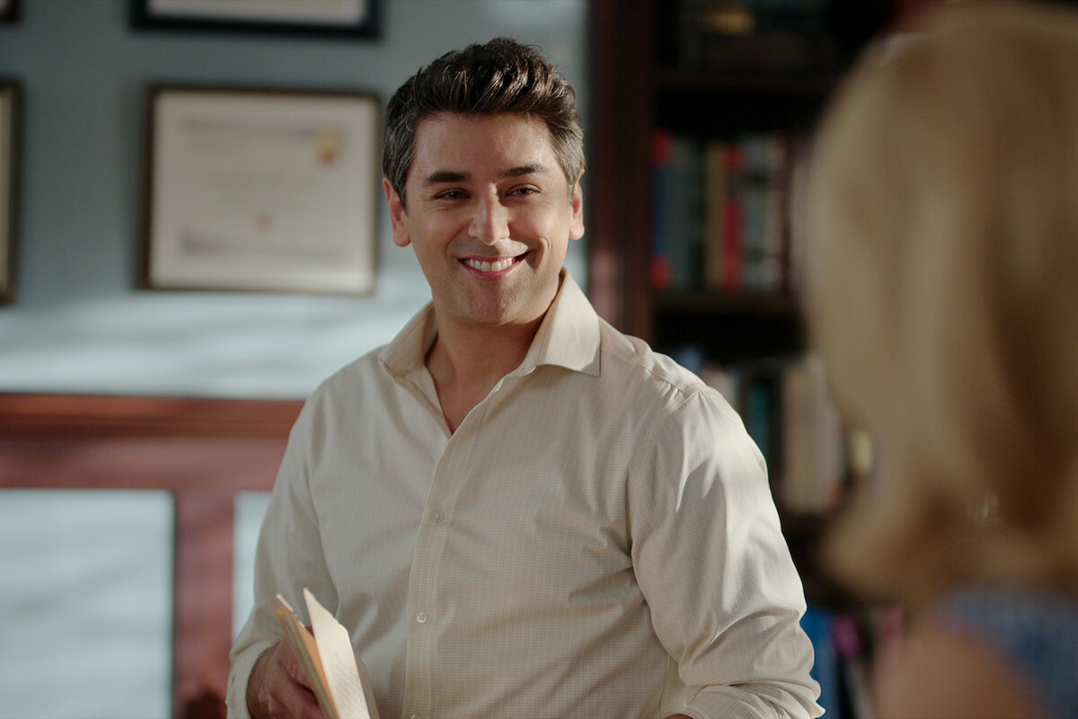 Mark Ghanimé as Dr. Cameron Hayek smiles in a doctor’s office in Season 5 of ‘Virgin River.’