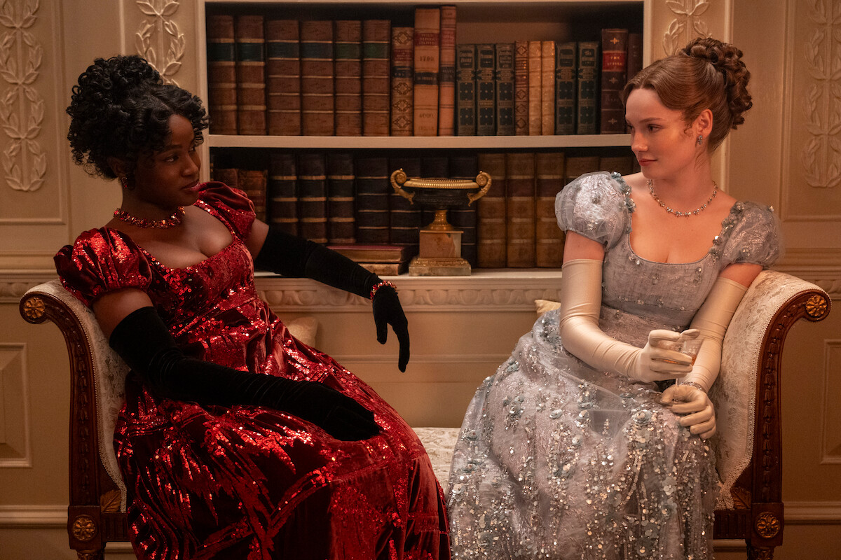 Two women in elegant Regency-era gowns and gloves sit on a vintage sofa in a lavish library, books and a gold trophy on shelves behind them, engaging in conversation.
