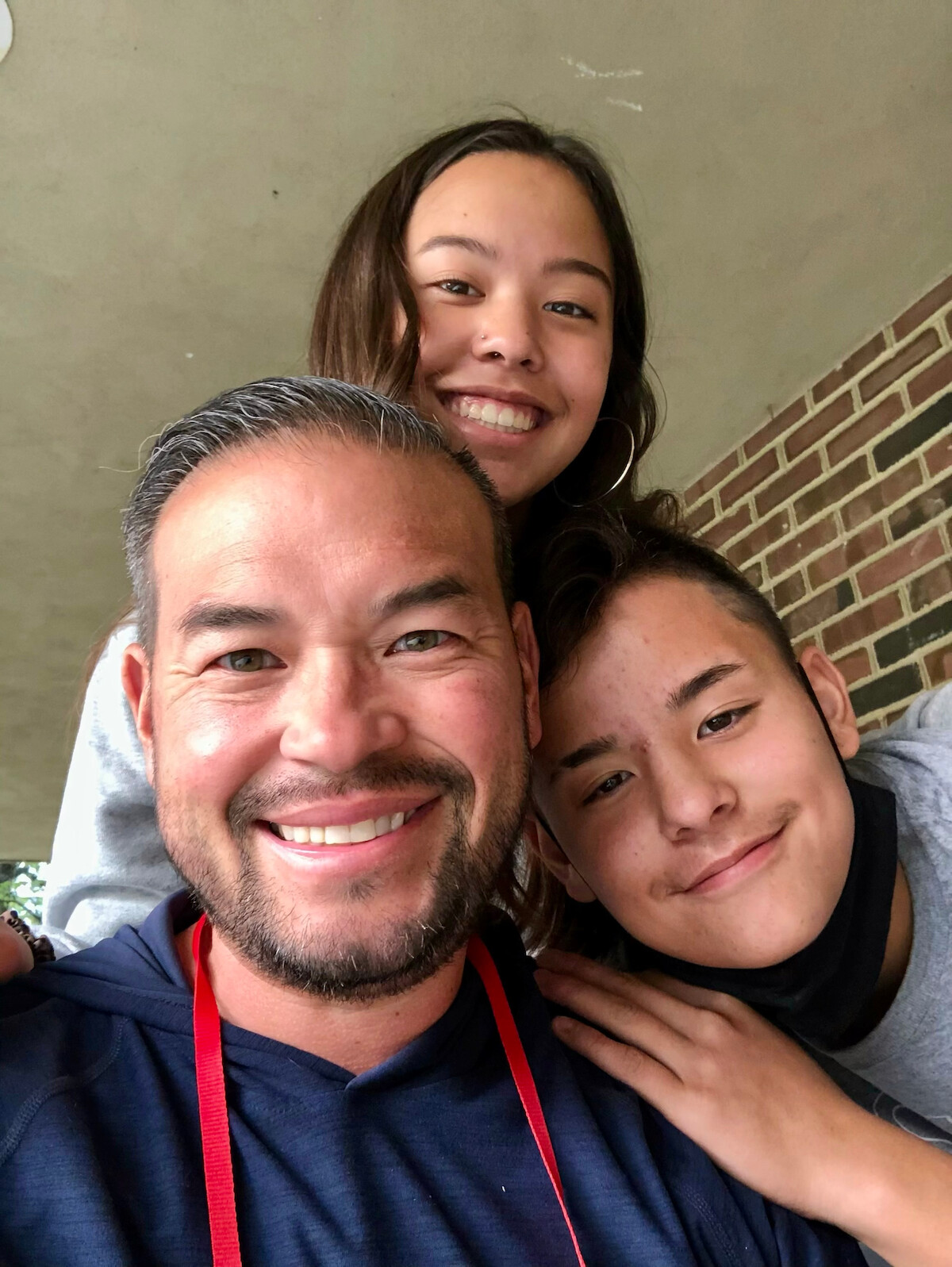 Three people smiling and posing closely together for a selfie outdoors, with a brick wall and ceiling in the background. They appear happy and relaxed.