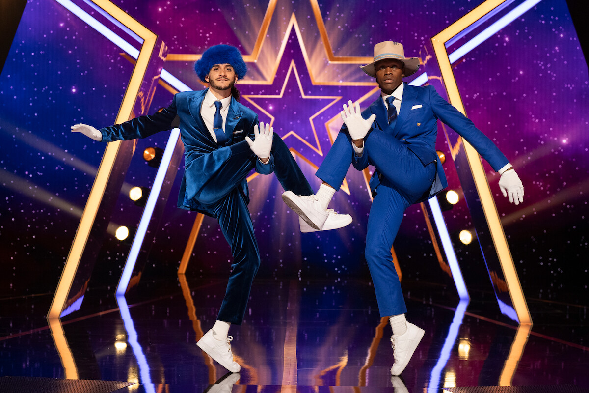 Kidd & Midas in blue suits, hats, and white gloves strike a synchronized dance pose on a shiny stage with star-themed purple and yellow lighting effects in the background.