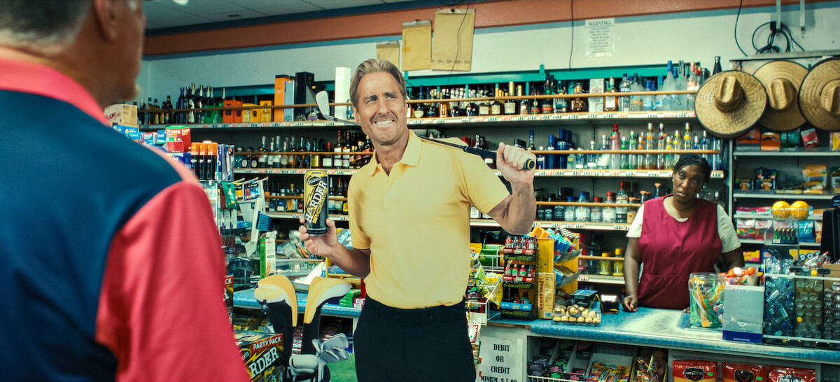 Man in yellow shirt holding a drink and gesturing in a colorful convenience store, another man in red shirt foreground, and store clerk behind counter. Shelves are stocked with snacks, drinks, and sombreros on wall.