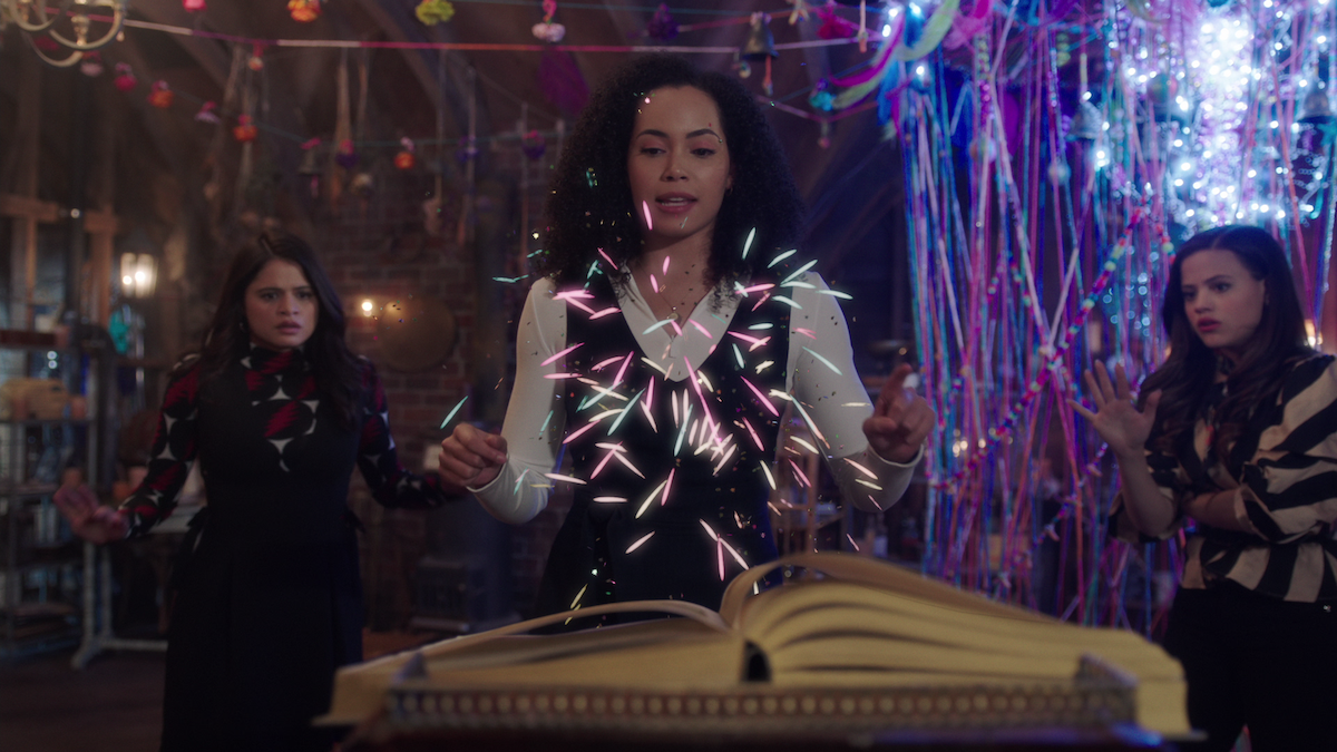 Three women in a decorated, magical room; one stands at an open book casting colorful sparks, while the others watch and gesture with concern. The environment is mystical with hanging lights and streamers.