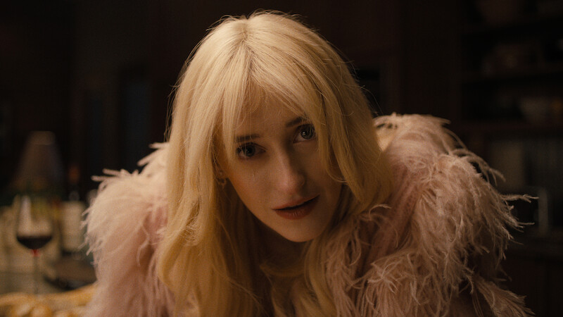 A woman with long blond hair and a light pink feathered jacket leans forward indoors at a table, with a glass of red wine in the background, warm lighting and a cozy, dimly lit environment.