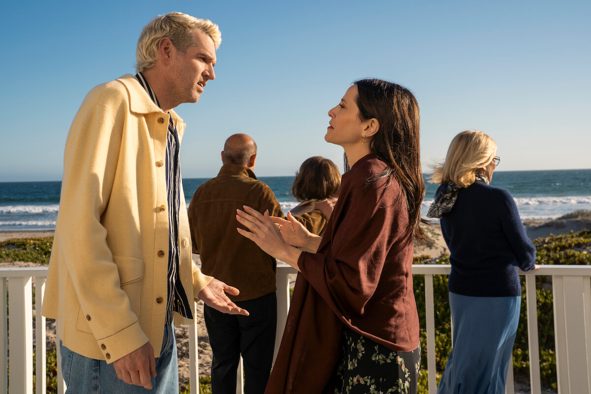 Two people in conversation on a sunny beachside deck, with three others looking out at the ocean, surrounded by greenery and blue sky.