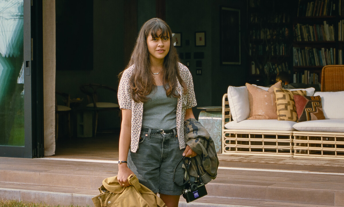Young woman with long hair stands outside a cozy house holding jackets, with a relaxed expression. The background includes a cushioned outdoor sofa, bookshelves, and soft natural lighting, creating a calm, inviting atmosphere.