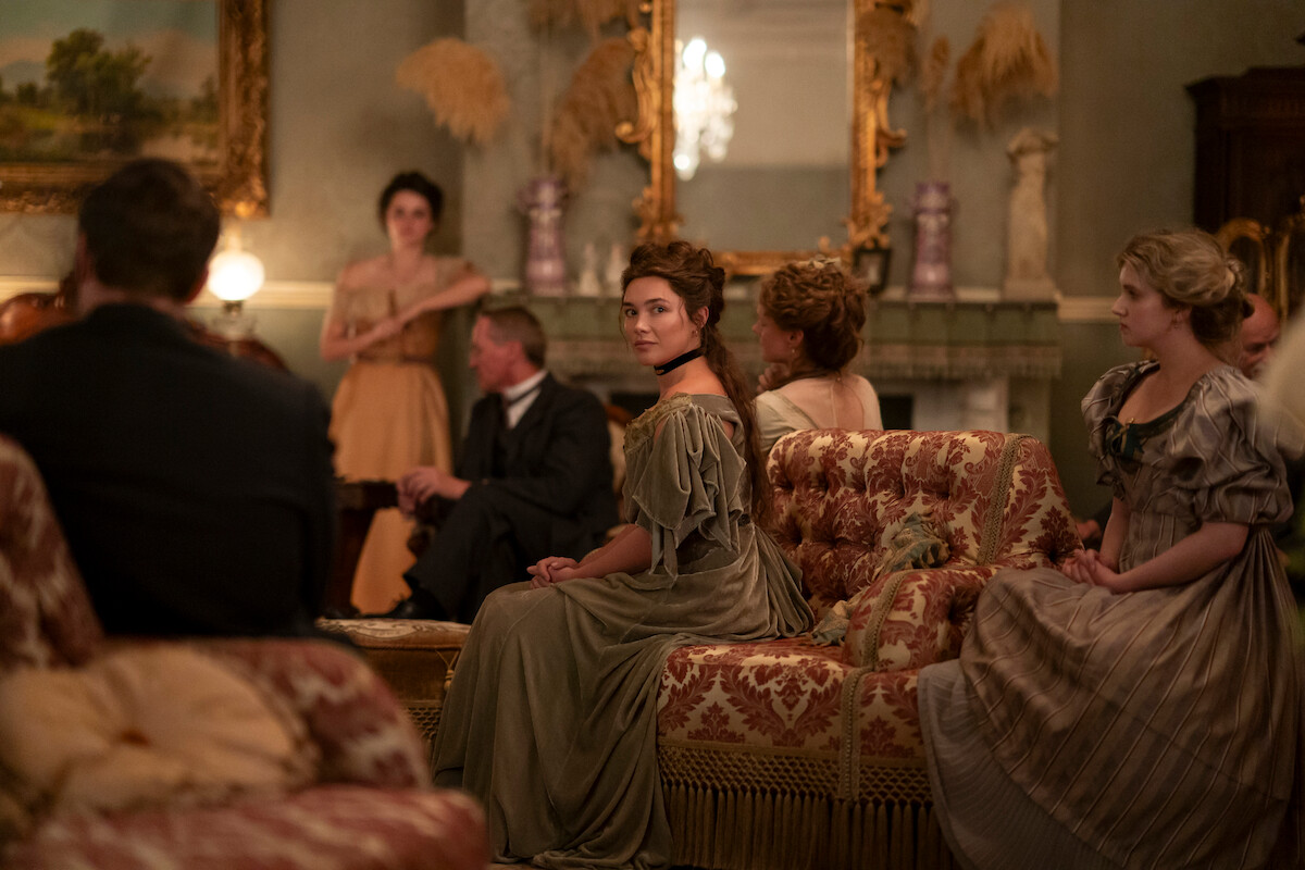 A group of people in period clothing sit in an ornate, vintage living room with elegant furniture, antique decorations, and a large mirror above the fireplace, engaged in conversation or listening.