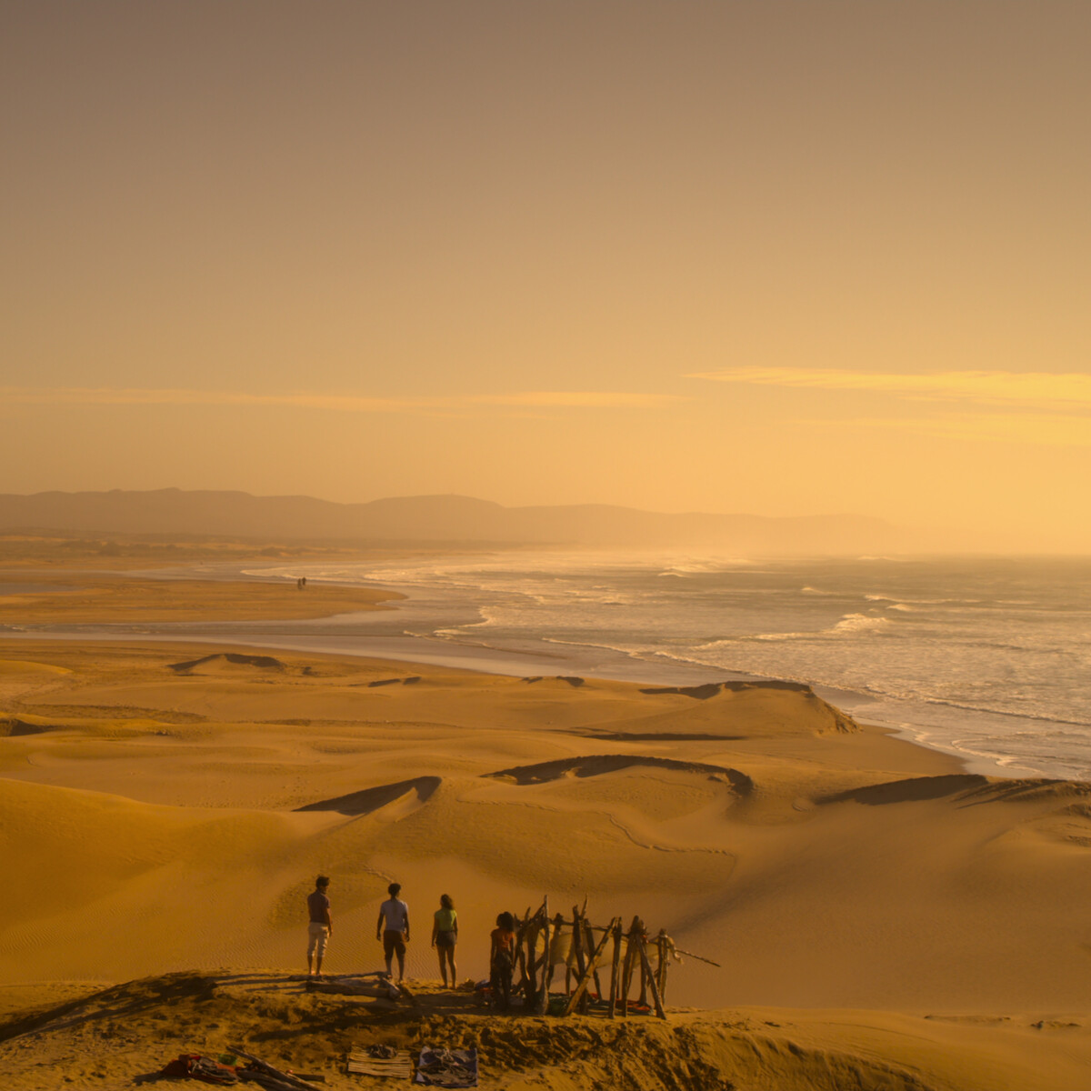 Group of people standing near a makeshift wooden structure on sandy dunes overlooking a vast beach and ocean at sunset, with distant hills under a golden sky.