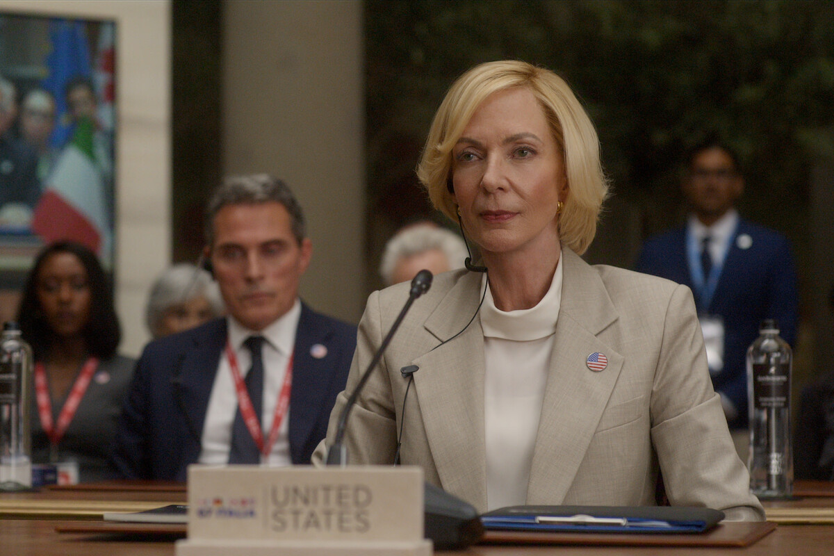 A woman in a light suit sits at a conference table with a United States nameplate and microphone, surrounded by delegates and flags, in a formal international meeting setting.