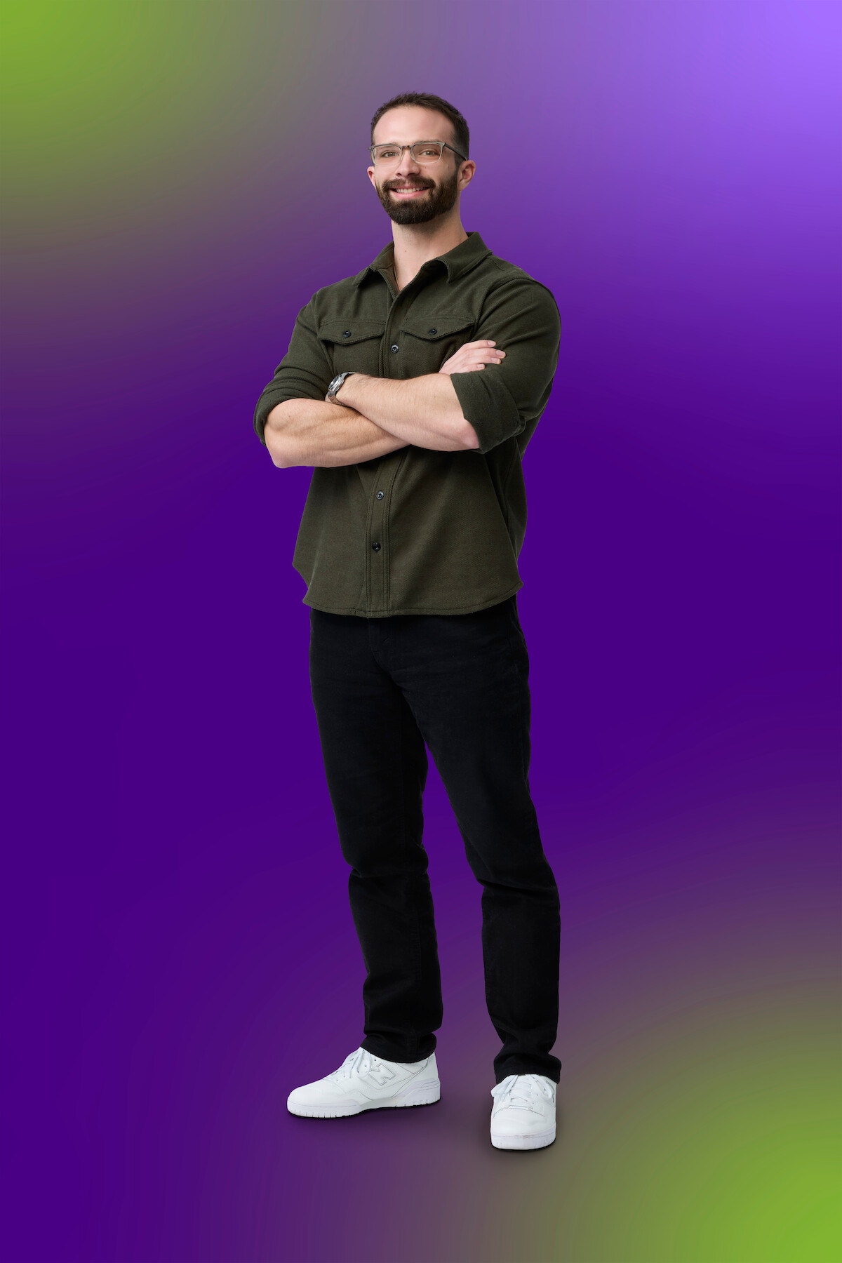 Man with glasses, beard, and short hair stands confidently with arms crossed, wearing a green shirt, black pants, and white sneakers, on a vibrant purple and green gradient background.