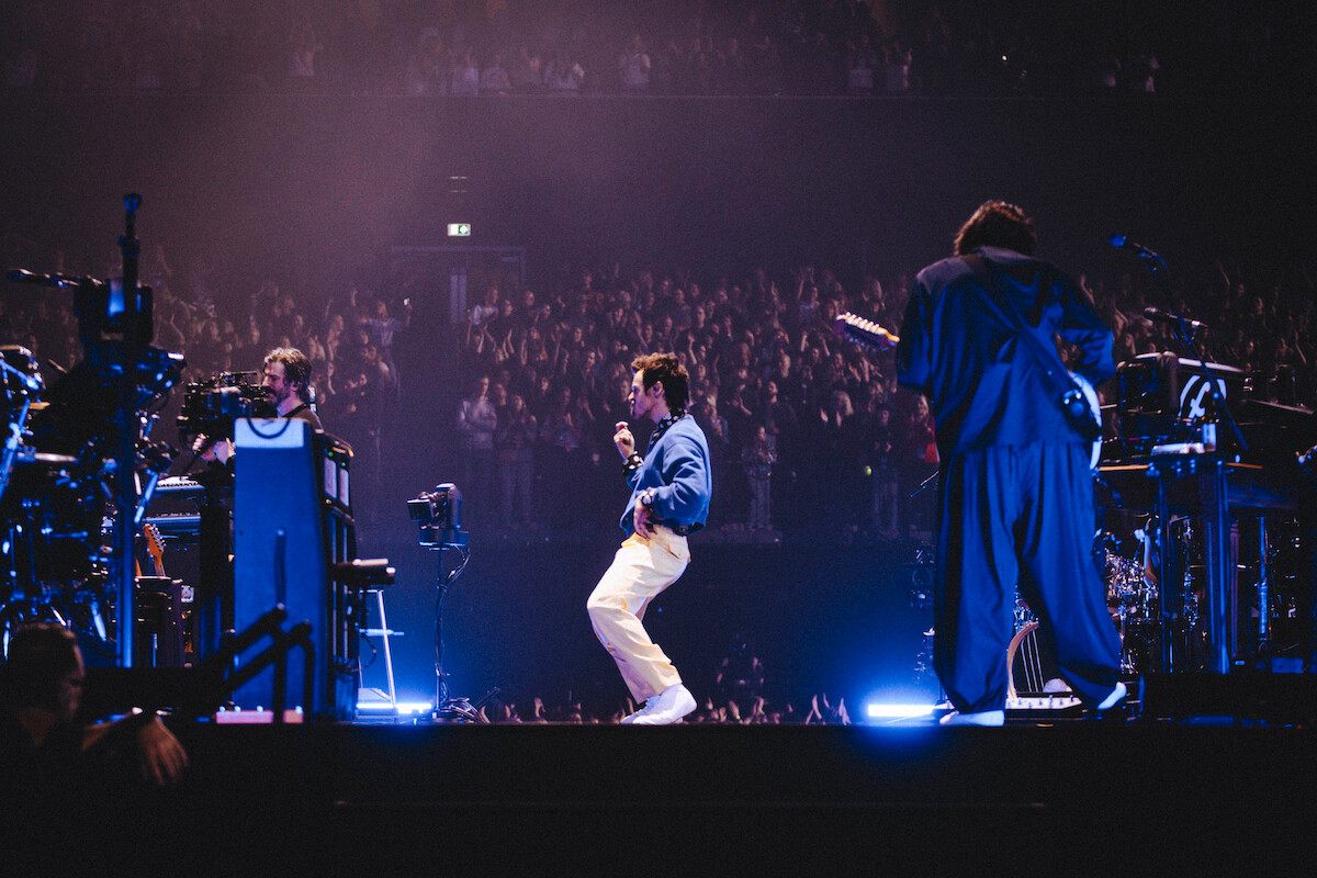 Harry Styles performing on stage with instruments, colorful stage lighting, and a large cheering audience in a dark concert venue.