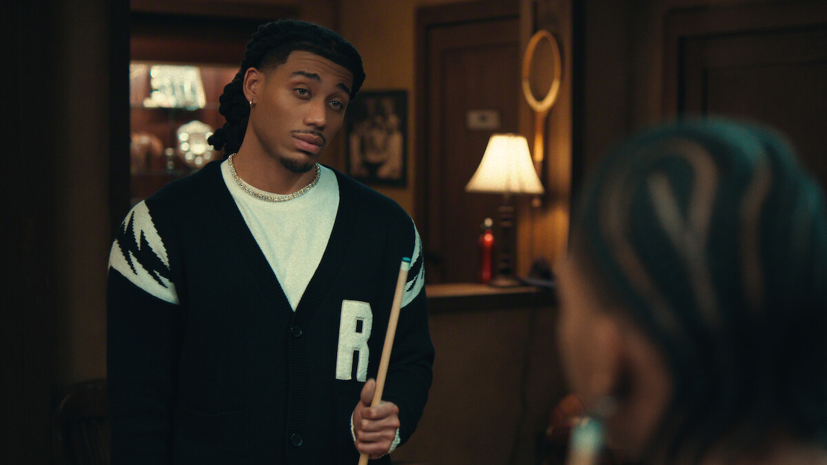 A man holding a pool cue stands in a warmly lit room, facing another person. There is a lamp on a table in the background, creating a cozy indoor environment with wood-paneled walls and decor.