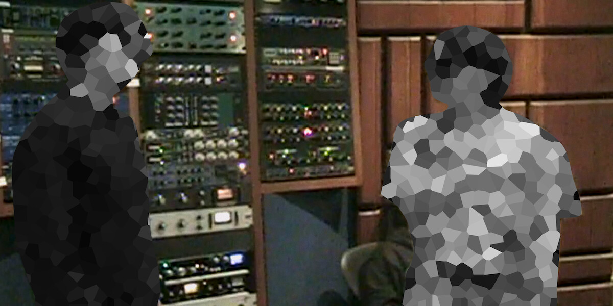 Two people in a recording studio stand near racks of audio equipment and mixers, surrounded by soundproofed walls and acoustic paneling.