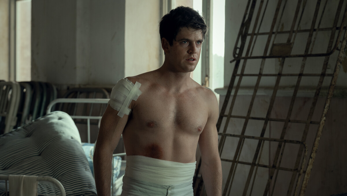 Shirtless young man with bandages on his shoulder and torso stands in a dimly lit, sparse room with metal bed frames and striped bedding, appearing to be in a hospital or infirmary setting.