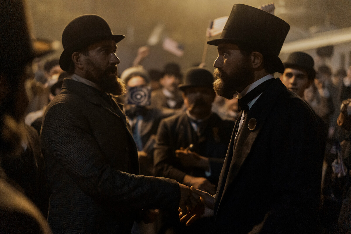 Two men in 19th-century attire shake hands in a dim, crowded outdoor setting, surrounded by onlookers with hats and flags, creating a formal, historical, and tense atmosphere.