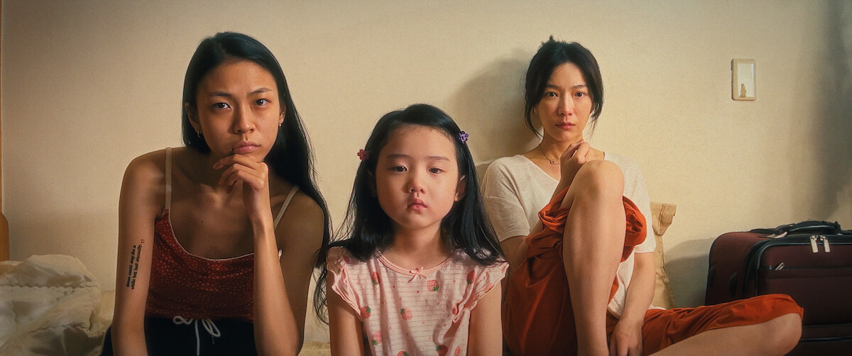 Three women sit closely on a bed in a softly lit room with neutral walls, appearing thoughtful and serious. There is a suitcase in the background. The overall mood is tense and contemplative.