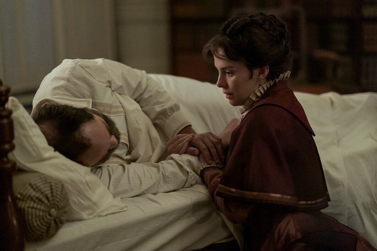 A woman in period clothing sits by a man's bedside, holding his hand as he lies in bed, appearing ill. The room is softly lit with a somber, intimate atmosphere, suggesting concern and care in a historical setting.