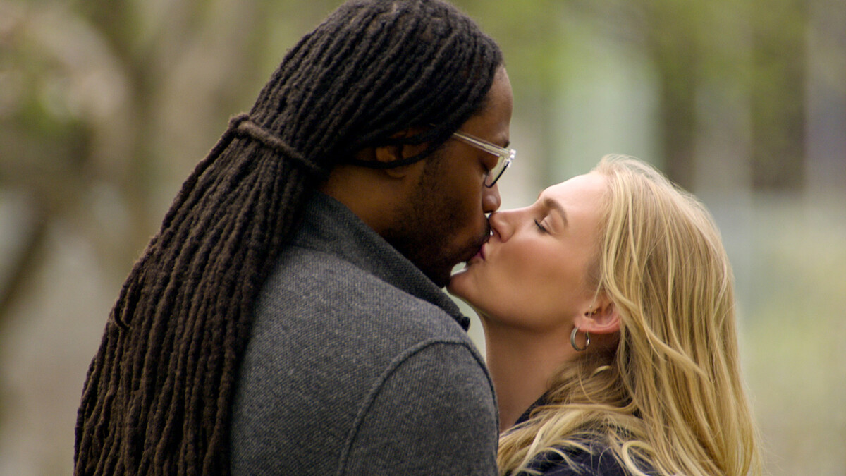 A couple sharing a kiss outdoors, with greenery and blurred trees in the background, suggesting a park or natural setting.