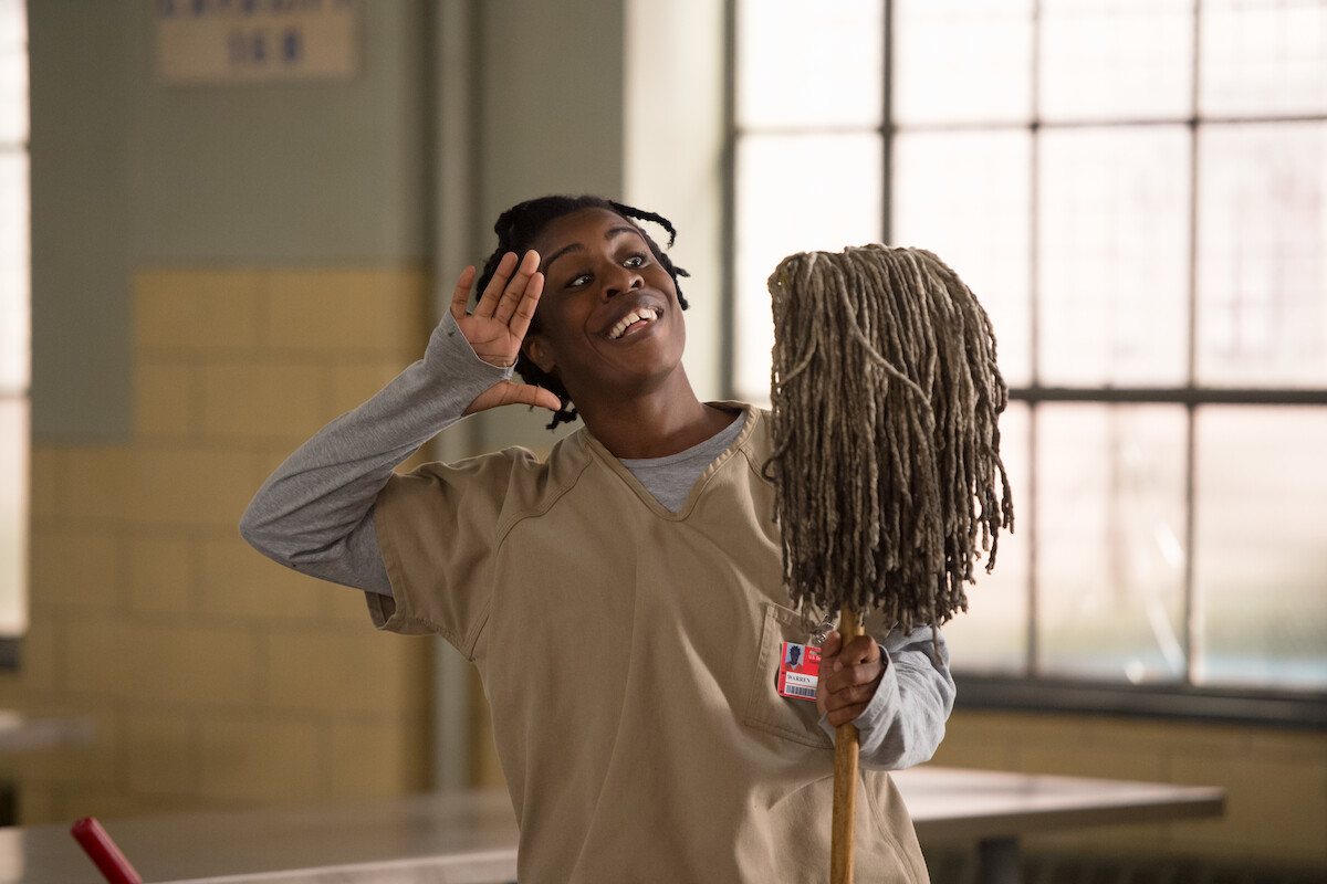 Uzo Aduba as Suzanne waves wide-eyed while smiling and holding a mop in Season 3 of ‘Orange Is the New Black.‘