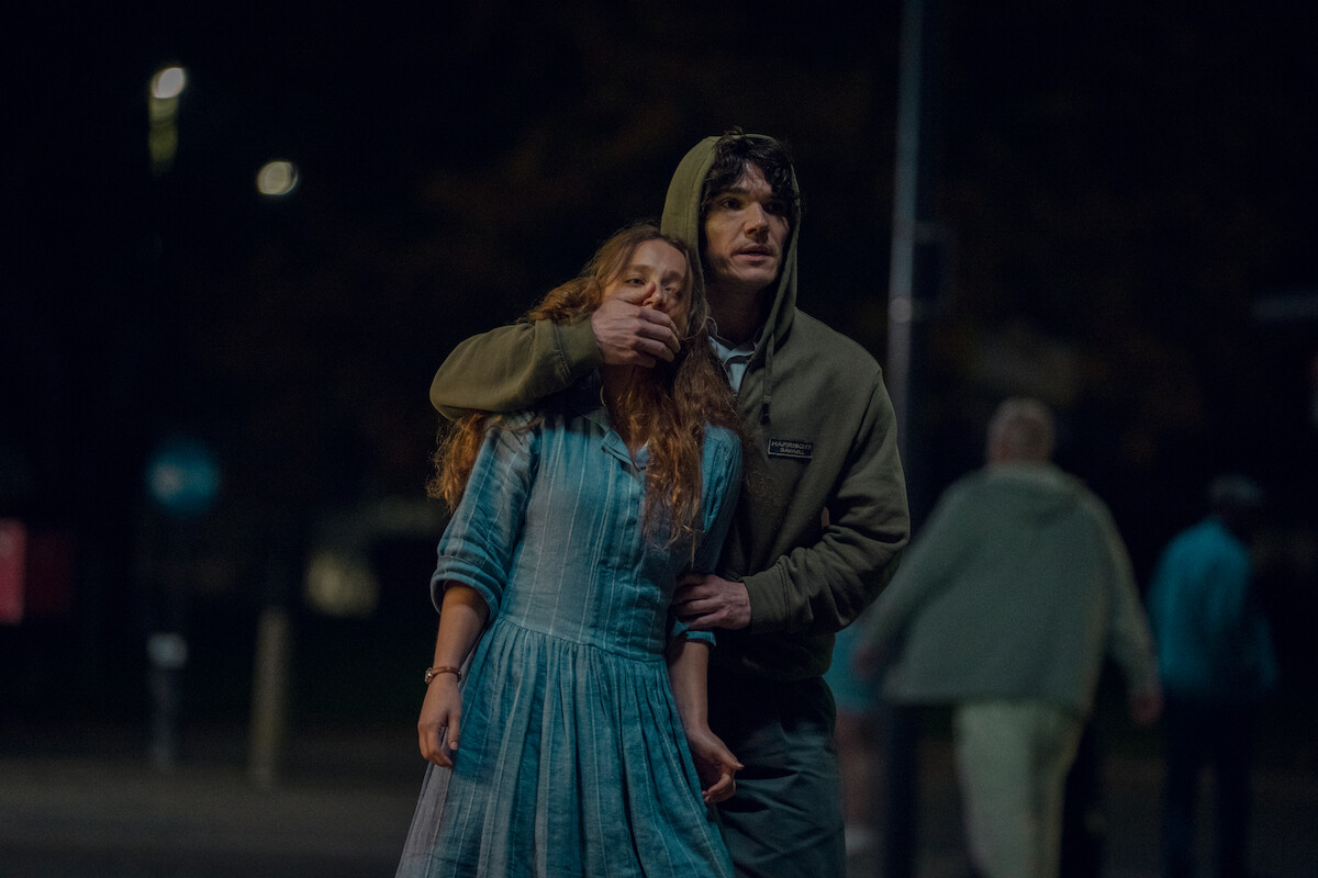 A man in a hoodie covers a woman's mouth and restrains her in a dimly lit outdoor area at night, with several people walking blurred in the background.