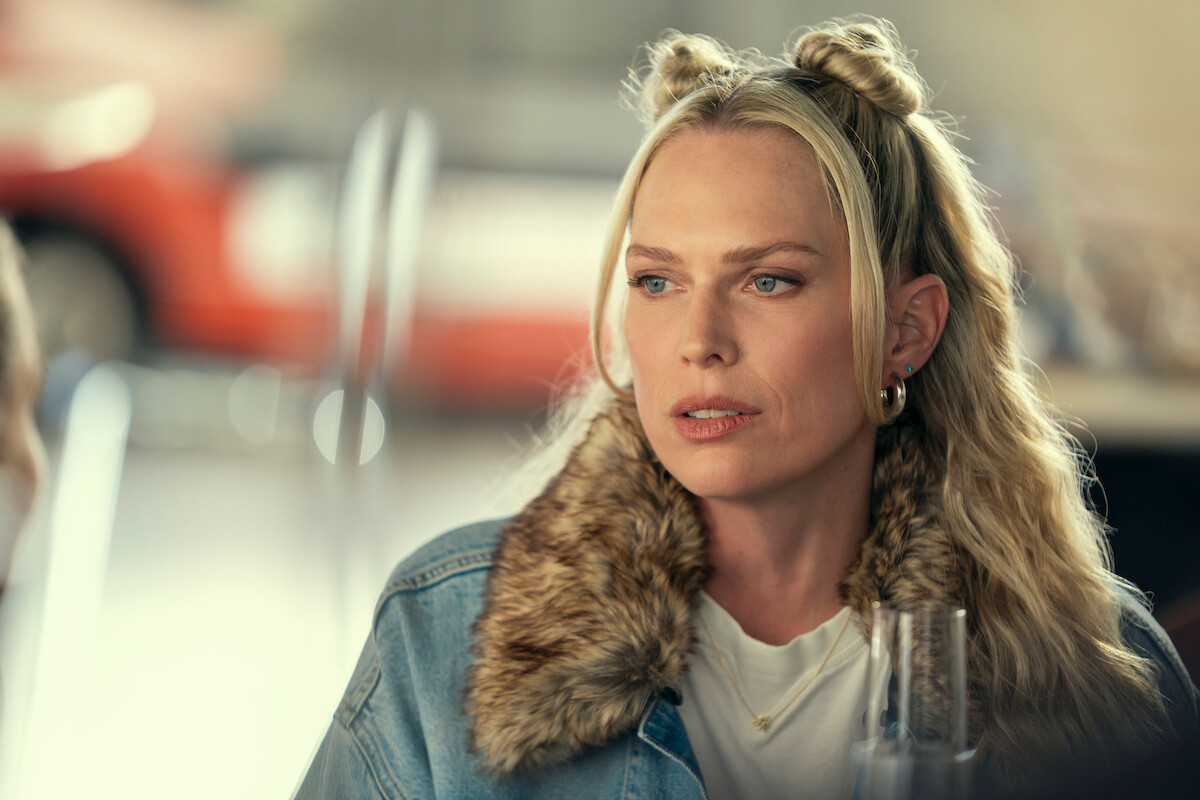 Woman with blonde hair in space buns, wearing a denim jacket with faux fur collar and hoop earrings, sitting indoors at a table in a softly lit environment, with blurred background suggesting a casual public setting.