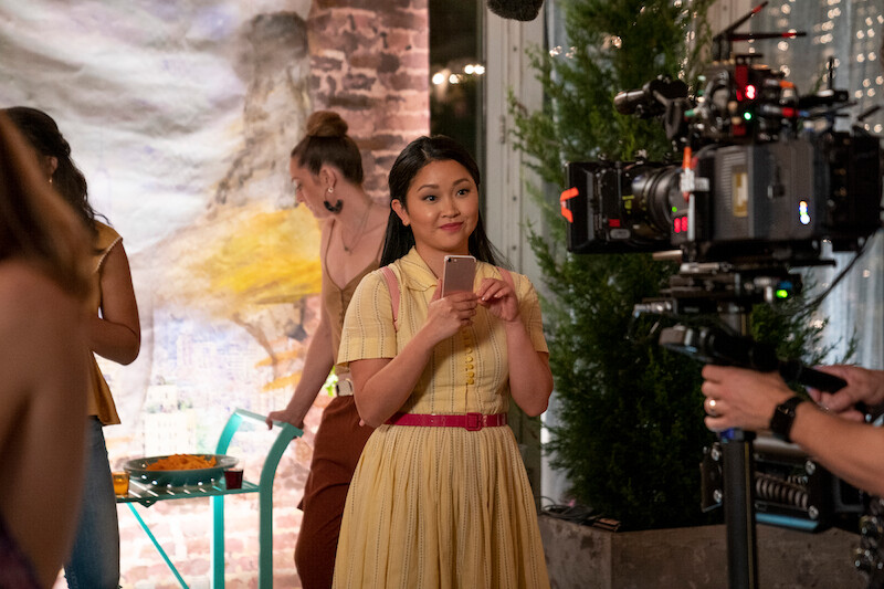Lana Condor as Lara Jean standing in front of a camera in a yellow dress holding a phone.