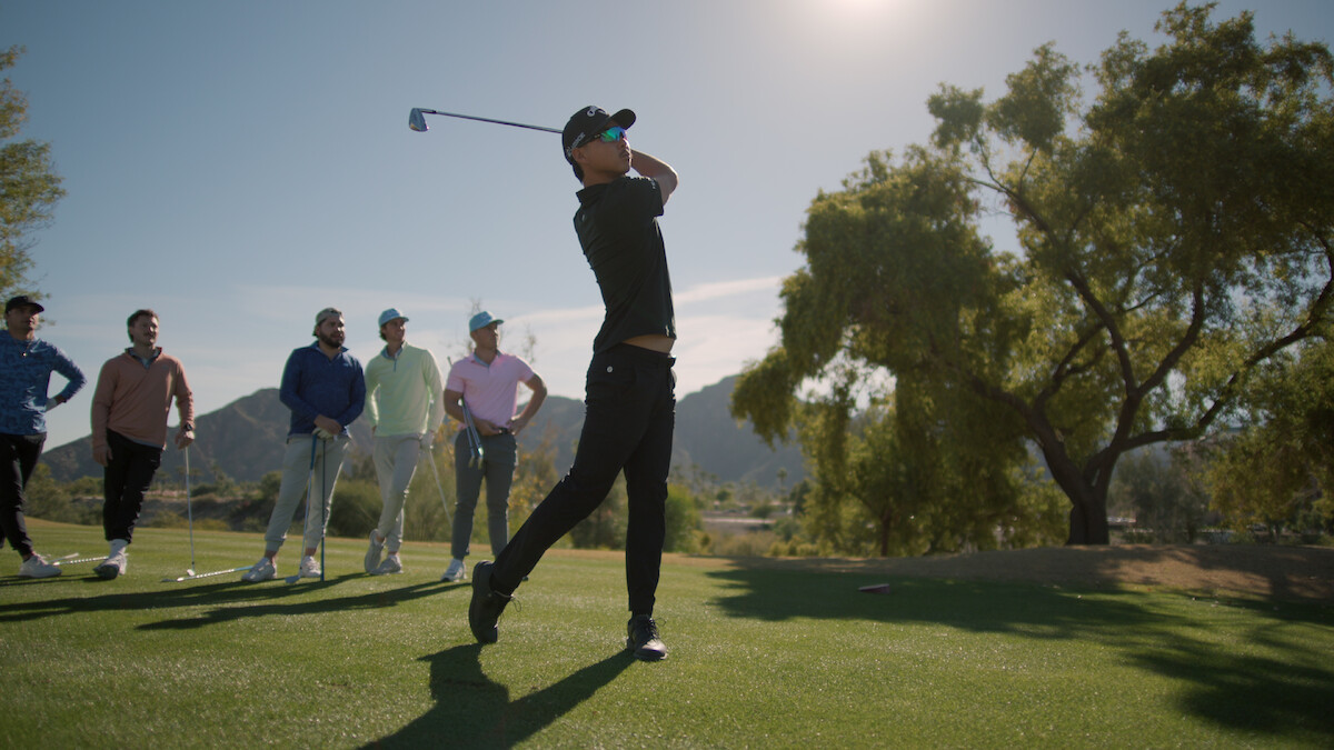 Min Woo Lee takes a swing with his golf club with onlookers behind him.